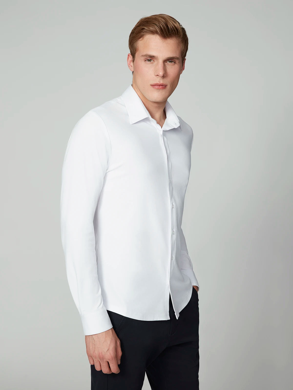 A man with short, light brown hair wears the PROJEK RAW MENS 24/7 BUTTON-UP KNIT SHIRT and dark pants, standing against a plain light gray background with one hand in his pocket.