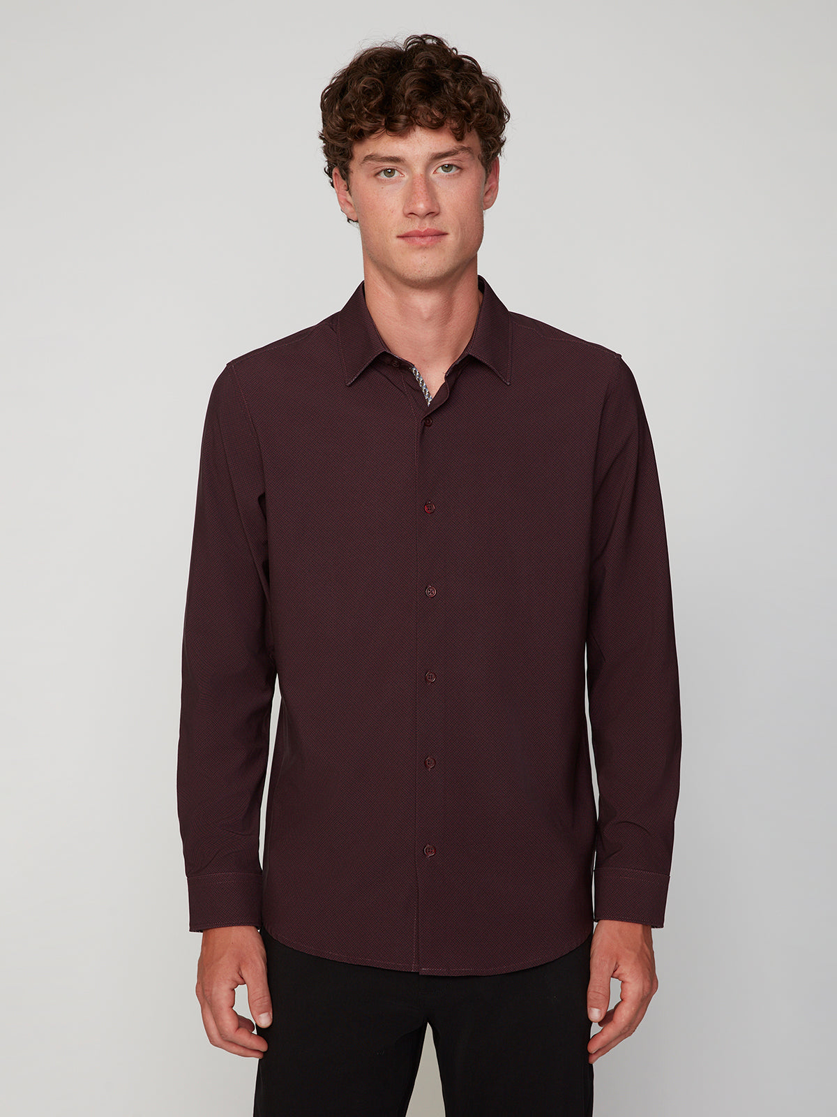 A young man with curly brown hair wears the PROJEK RAW MENS LONG SLEEVE 4WAY STRETCH TWILL SHIRT in dark maroon and black pants, standing against a plain, light gray background.