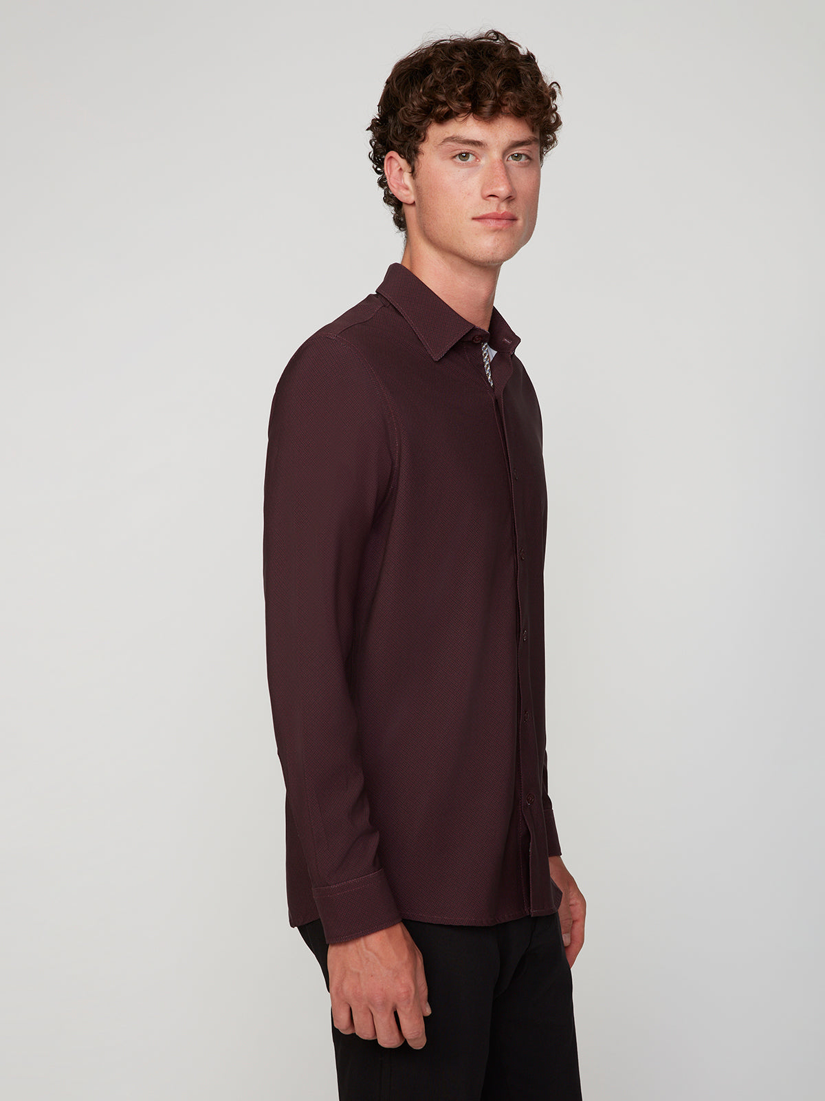 A young man with curly brown hair models the PROJEK RAW MENS LONG SLEEVE 4WAY STRETCH TWILL SHIRT in burgundy and black pants, standing sideways and looking at the camera against a plain light gray background.