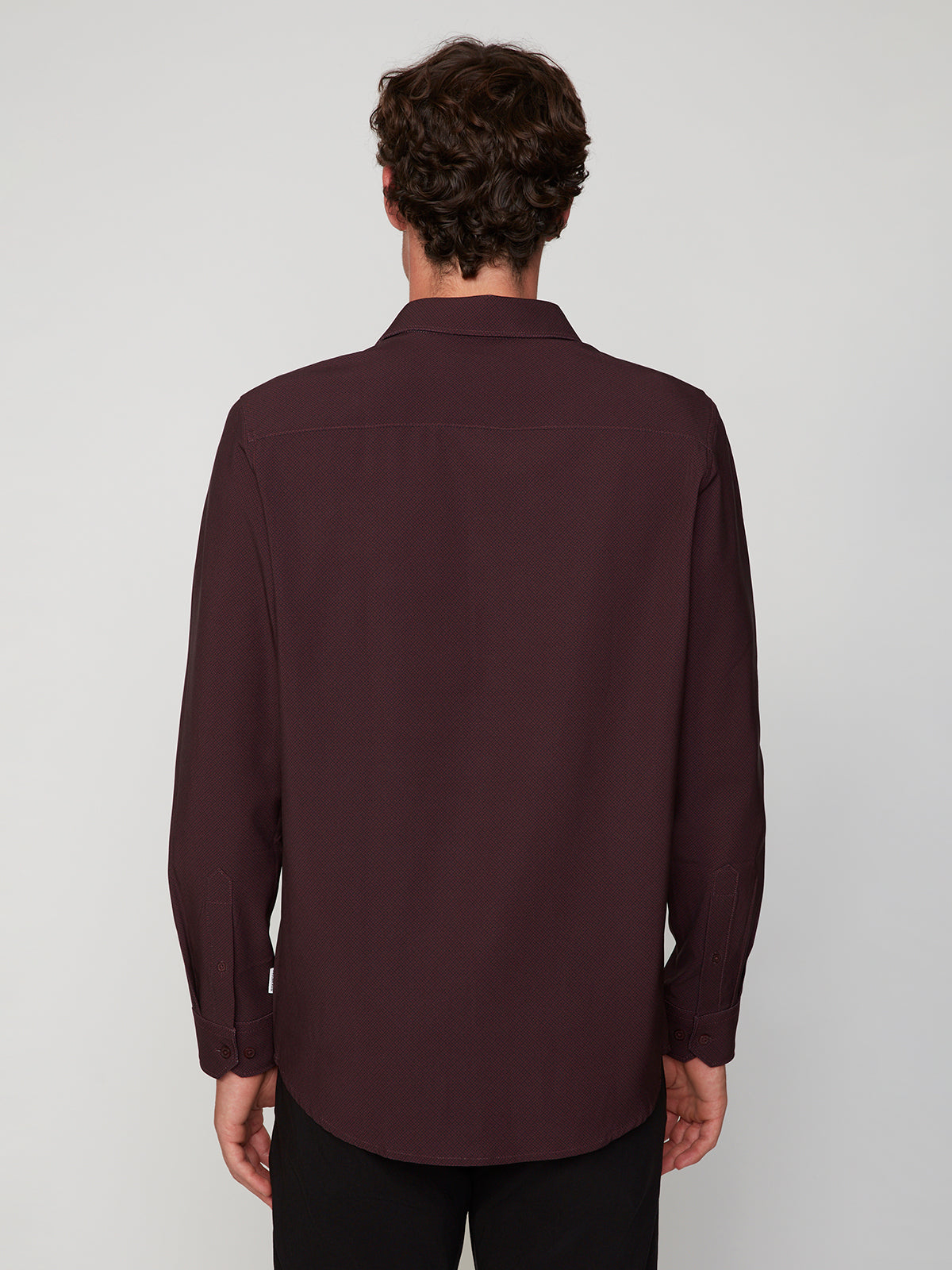 A man with short, curly brown hair is seen from behind wearing the PROJEK RAW MENS LONG SLEEVE 4WAY STRETCH TWILL SHIRT in dark burgundy and black pants, set against a plain light gray background.