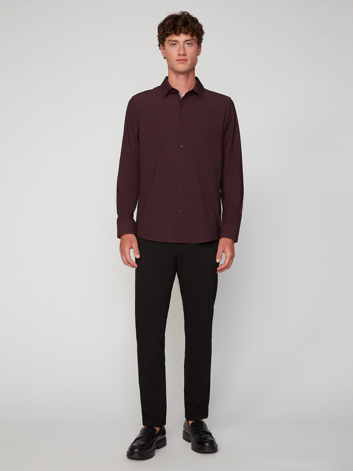 A young man with curly brown hair stands facing forward, wearing the PROJEK RAW MENS LONG SLEEVE 4WAY STRETCH TWILL SHIRT in dark maroon, paired with black pants and shoes, against a plain light gray background.